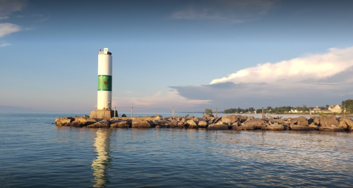 A lighthouse stands on a rocky pier, reflecting in calm waters under a clear sky with distant land visible.