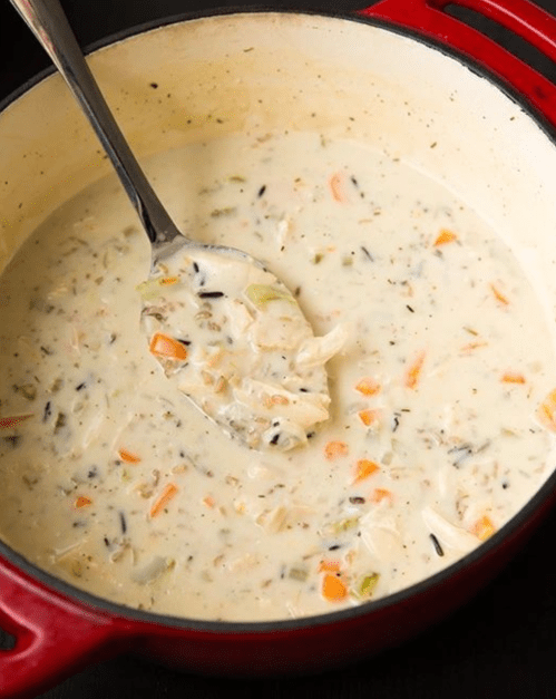 A pot of creamy chicken and wild rice soup with vegetables, featuring a spoon resting inside.