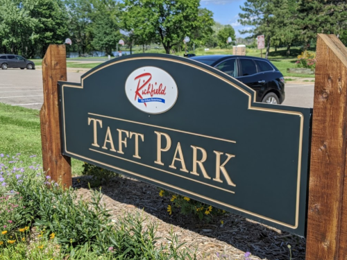 Sign for Taft Park in Richfield, surrounded by greenery and flowers, with a parking lot and trees in the background.