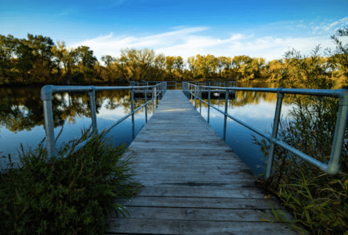 A wooden dock extends into a calm lake, surrounded by trees and reflecting the blue sky.