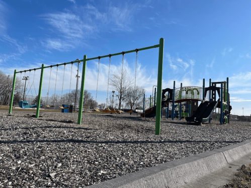 A playground with swings and a slide, set against a clear blue sky and trees in the background.