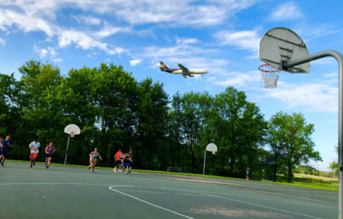 A basketball court with players running, while an airplane flies overhead against a blue sky and green trees.