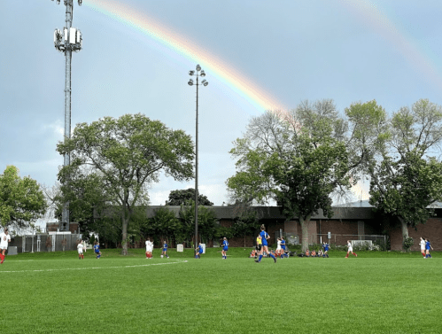 A soccer game in progress on a green field, with a rainbow arching over the scene and trees in the background.