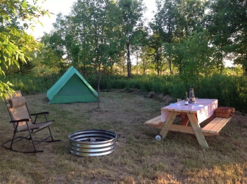 A cozy campsite with a green tent, picnic table, rocking chair, and fire pit surrounded by trees.