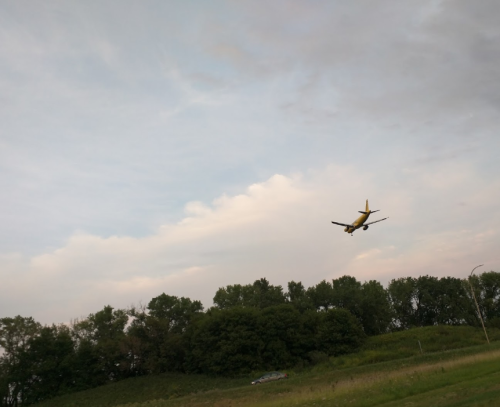 An airplane approaches for landing over a green landscape under a cloudy sky.