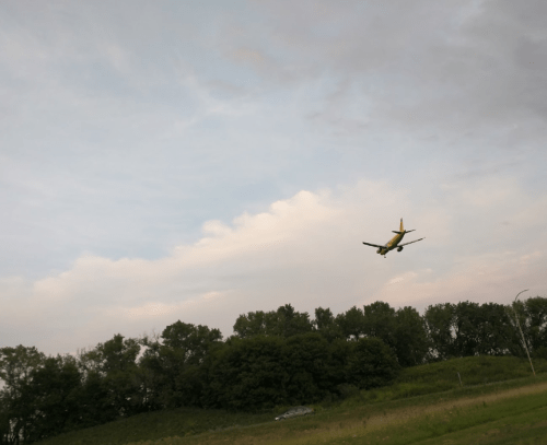 An airplane approaches for landing over a green landscape under a cloudy sky.