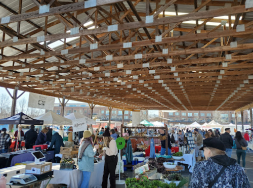 A bustling farmers market under a wooden pavilion, with vendors and shoppers among various stalls and produce.
