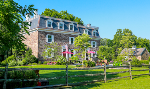A charming stone house with a manicured garden, surrounded by trees and a wooden fence, under a clear blue sky.