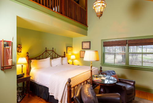 Cozy bedroom with a large bed, warm lighting, and a sitting area, featuring green walls and a window view.