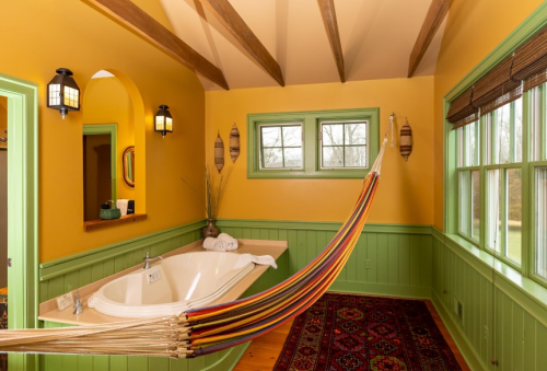 A cozy bathroom with a yellow and green color scheme, featuring a hammock, a bathtub, and large windows.