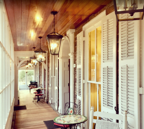 A cozy porch with wooden floors, white shutters, and hanging lanterns, featuring small tables and chairs.