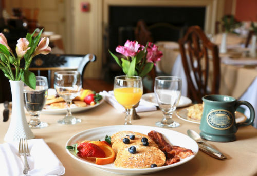A beautifully set breakfast table with pastries, bacon, fruit, and drinks, surrounded by flowers in vases.
