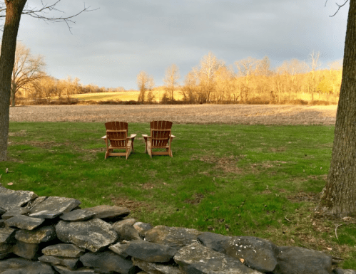 Two wooden chairs facing a grassy field, framed by trees and a stone wall, under a cloudy sky.