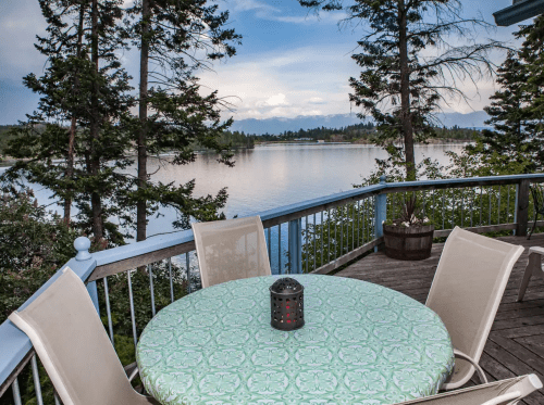 A serene lakeside view from a deck with a round table and chairs, surrounded by trees and mountains in the distance.