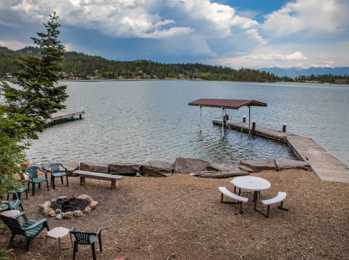 A serene lakeside view featuring a dock, seating area, and fire pit surrounded by trees and mountains.