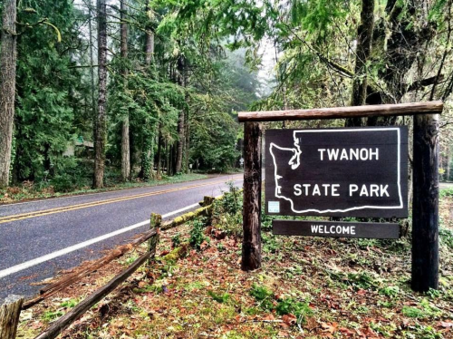 Welcome sign for Twanoh State Park, surrounded by lush green trees and a misty atmosphere along a quiet road.