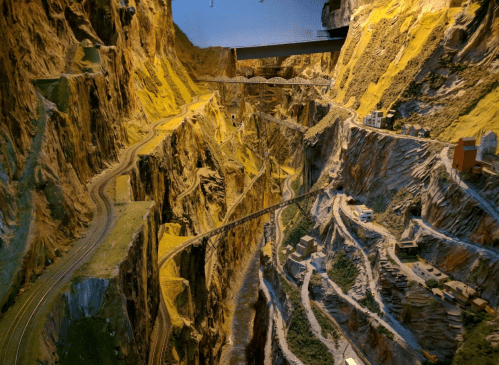 A detailed model of a mountainous landscape with winding train tracks and small buildings nestled in the rocky terrain.