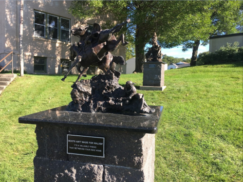 Two bronze sculptures on stone pedestals in a grassy area, depicting dynamic figures in motion.
