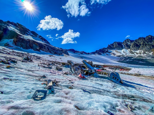 Wreckage of an aircraft on a snowy mountain landscape under a bright blue sky with scattered clouds.