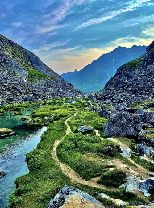 A serene mountain landscape with a winding path, lush greenery, and a clear stream under a colorful sky.