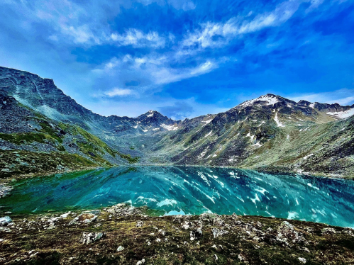 A serene mountain landscape with a clear blue lake reflecting the sky and surrounding peaks under a partly cloudy sky.