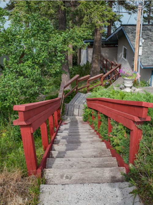 A wooden staircase with red railings descends through greenery, leading to a house in a forested area.