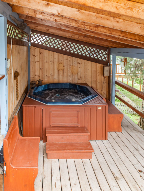 A wooden hot tub under a rustic roof, surrounded by wooden walls and a deck, with a bench nearby.