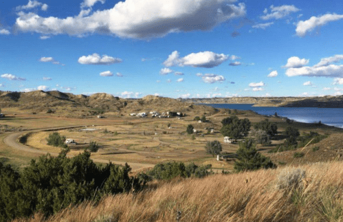 A scenic landscape featuring rolling hills, a lake, and scattered clouds in a blue sky. Green grass and trees are visible.