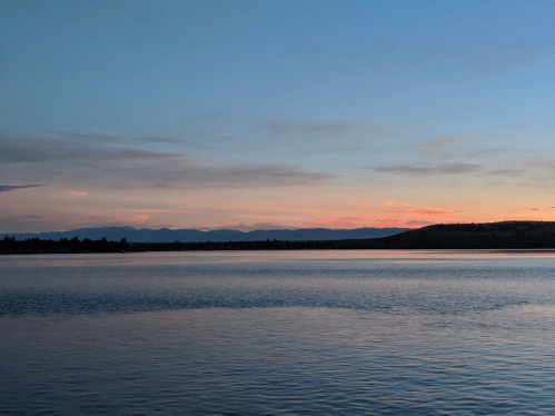 A serene lake at sunset, with soft pink and orange hues reflecting on the water and mountains in the background.
