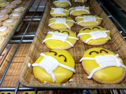 A tray of yellow smiley face cookies wearing white masks, arranged neatly in a bakery display.