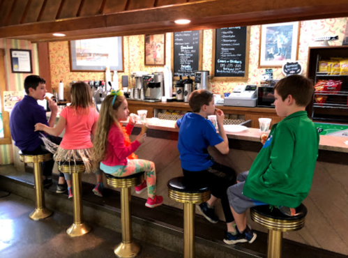 Five children sit at a retro-style counter, enjoying ice cream and drinks in a colorful, casual setting.