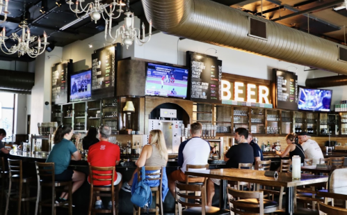 A lively bar interior with patrons seated at the counter, large screens showing sports, and a prominent "BEER" sign.