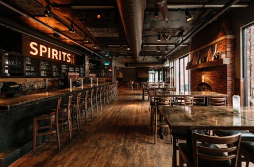 A spacious restaurant interior with wooden tables, chairs, and a bar featuring a "SPIRITS" sign. Large windows let in natural light.