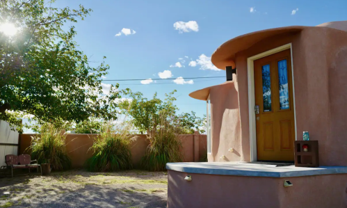 A round, adobe-style building with a wooden door, surrounded by greenery and a clear blue sky.
