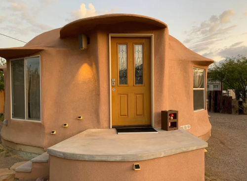 A unique, rounded adobe house with a bright yellow door and large windows, set against a sunset sky.
