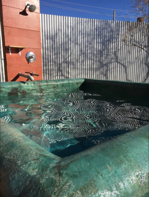 A green, rectangular hot tub with rippling water, surrounded by a metal fence and a clear blue sky.