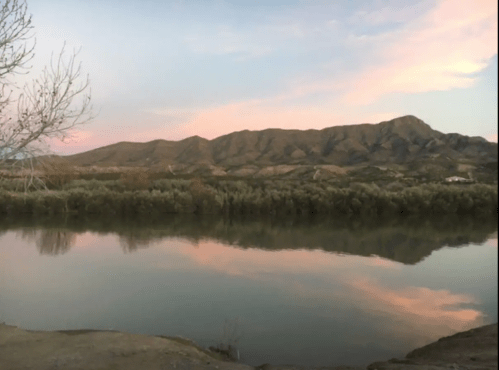 A serene landscape featuring a calm river reflecting pink skies and mountains in the background during sunset.