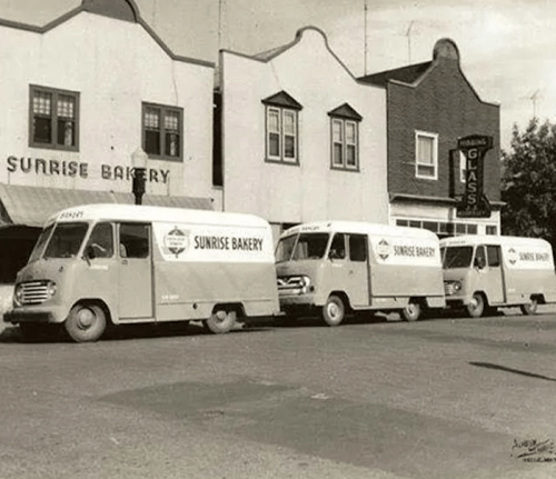 Vintage photo of bakery delivery vans parked outside Sunrise Bakery, with a storefront and nearby buildings in view.