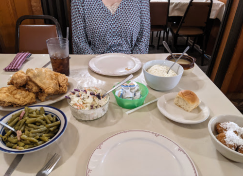 A table set with fried chicken, green beans, coleslaw, a roll, and dessert, with a person partially visible in the background.