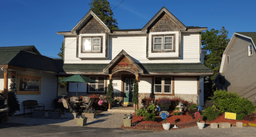 A charming two-story house with a green roof, surrounded by colorful flowers and outdoor seating.