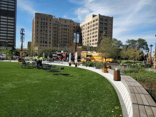 A green park area with benches, trees, and food trucks in an urban setting, surrounded by buildings under a clear sky.