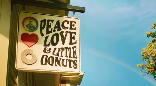 Sign reading "Peace Love & Little Donuts" with a heart and donut graphic against a blue sky.
