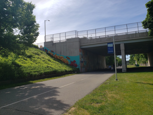 A pathway leads under a bridge with colorful murals on the side, surrounded by greenery and a clear blue sky.