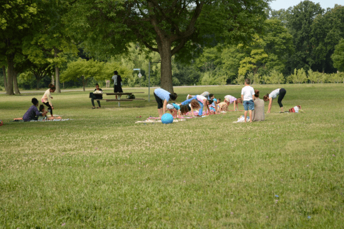 A group of children playing on a grassy field, with adults sitting nearby under trees on a sunny day.