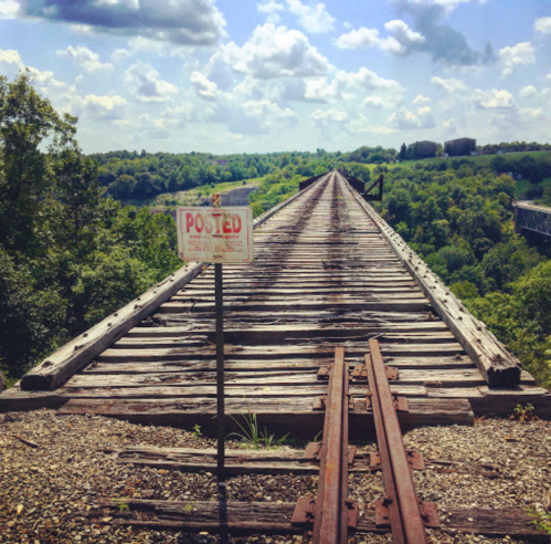 A wooden railway bridge stretches into the distance, surrounded by lush greenery and a blue sky with fluffy clouds.