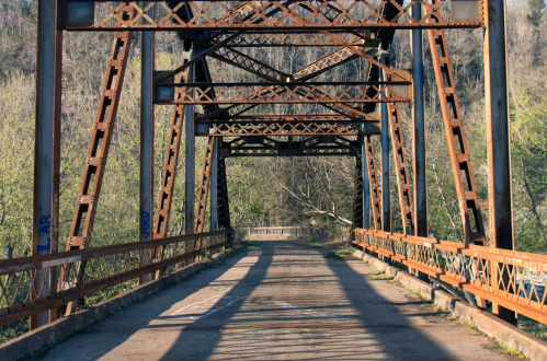 Rusty metal bridge with a clear path, surrounded by greenery and shadows, leading into the distance.
