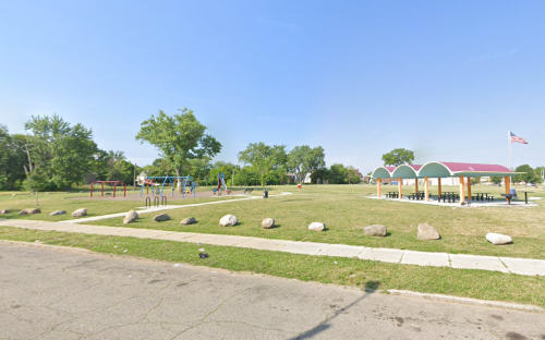 A sunny park featuring a playground, picnic area with a roof, and grassy space, bordered by a street and rocks.