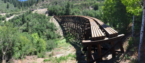 A wooden bridge arches over a lush green landscape, surrounded by trees and hills under a clear blue sky.