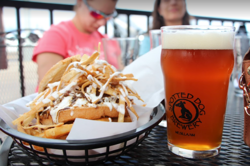 A plate of loaded fries topped with sauce next to a pint of amber beer on a table.