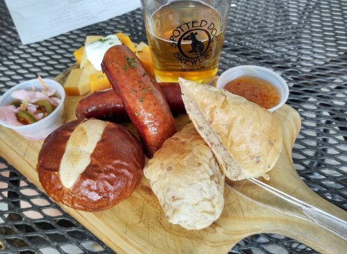 A wooden platter with assorted sausages, rolls, cheese, pickled vegetables, and a beer on a table.